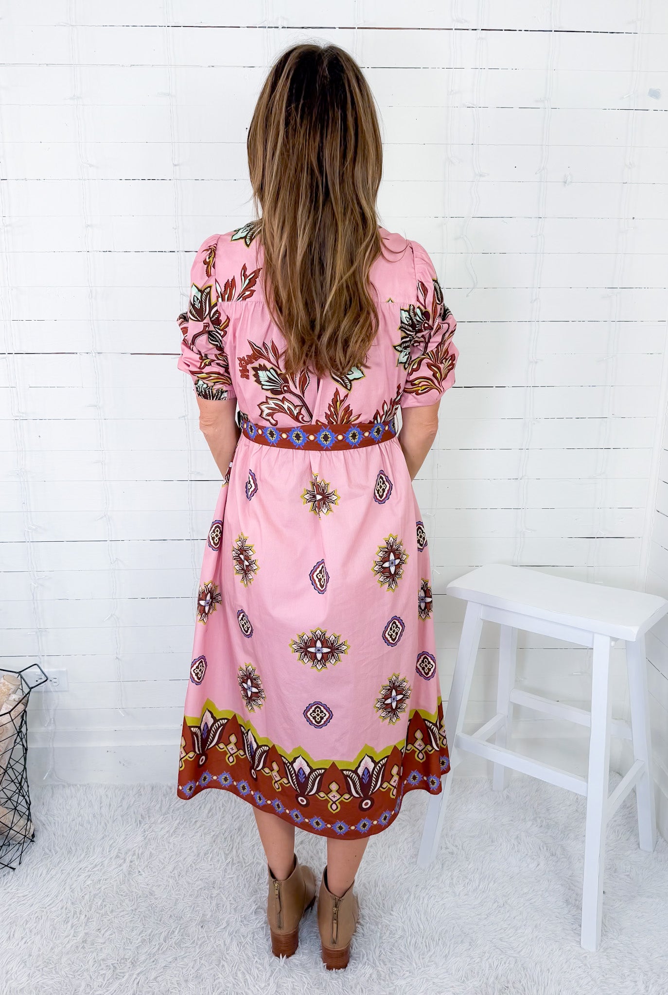 Woman wearing a pink dress with a pattern, standing against a white wooden panel wall.