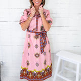 Woman wearing a pink dress with a pattern, standing against a white wooden panel background.