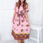 Woman wearing a pink patterned dress sitting on a white stool against a white wooden wall.