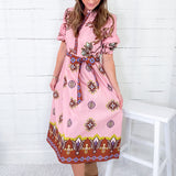 Woman wearing a pink patterned dress sitting on a white stool against a white wooden wall.