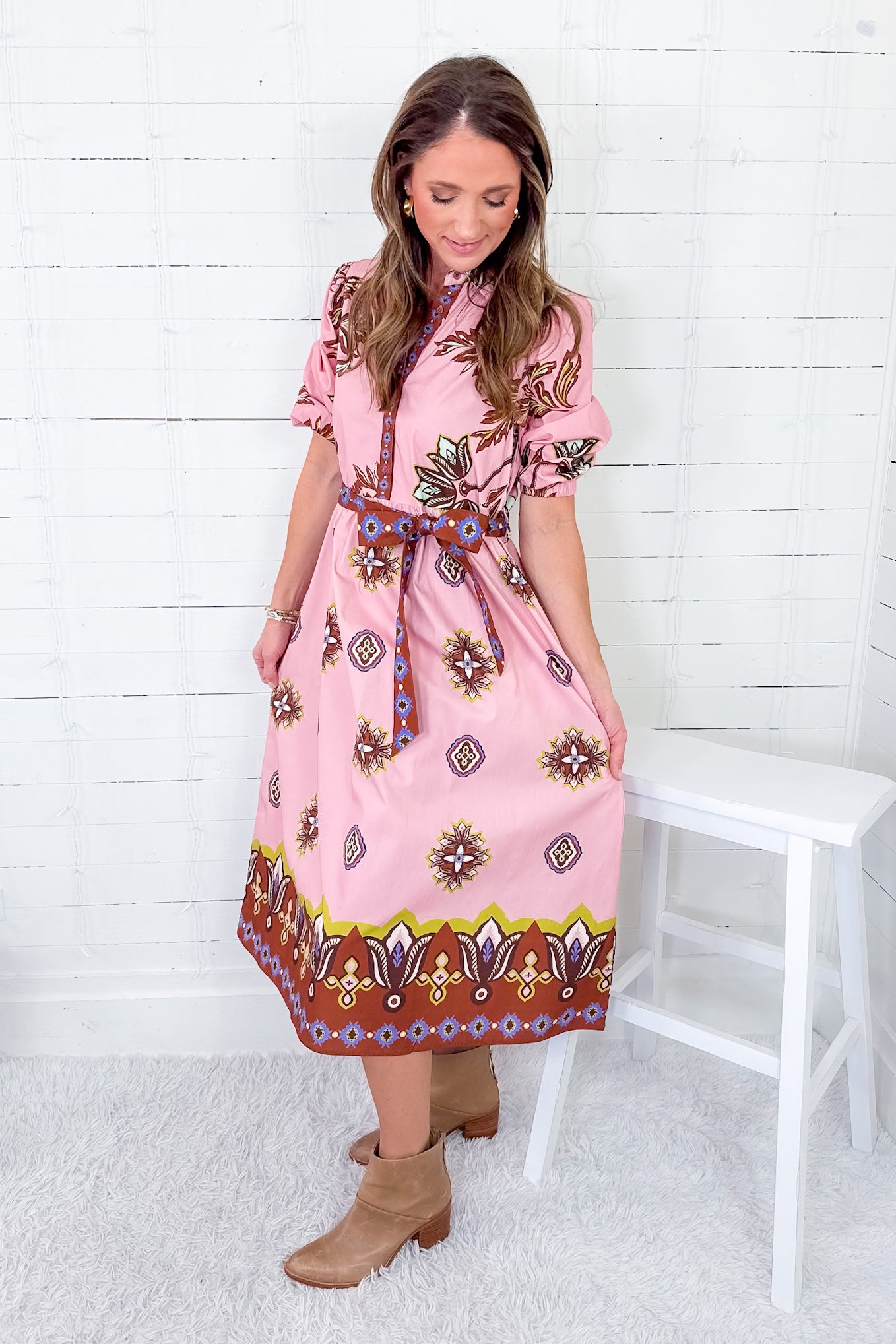 Woman wearing a pink patterned dress sitting on a white stool against a white wooden wall.