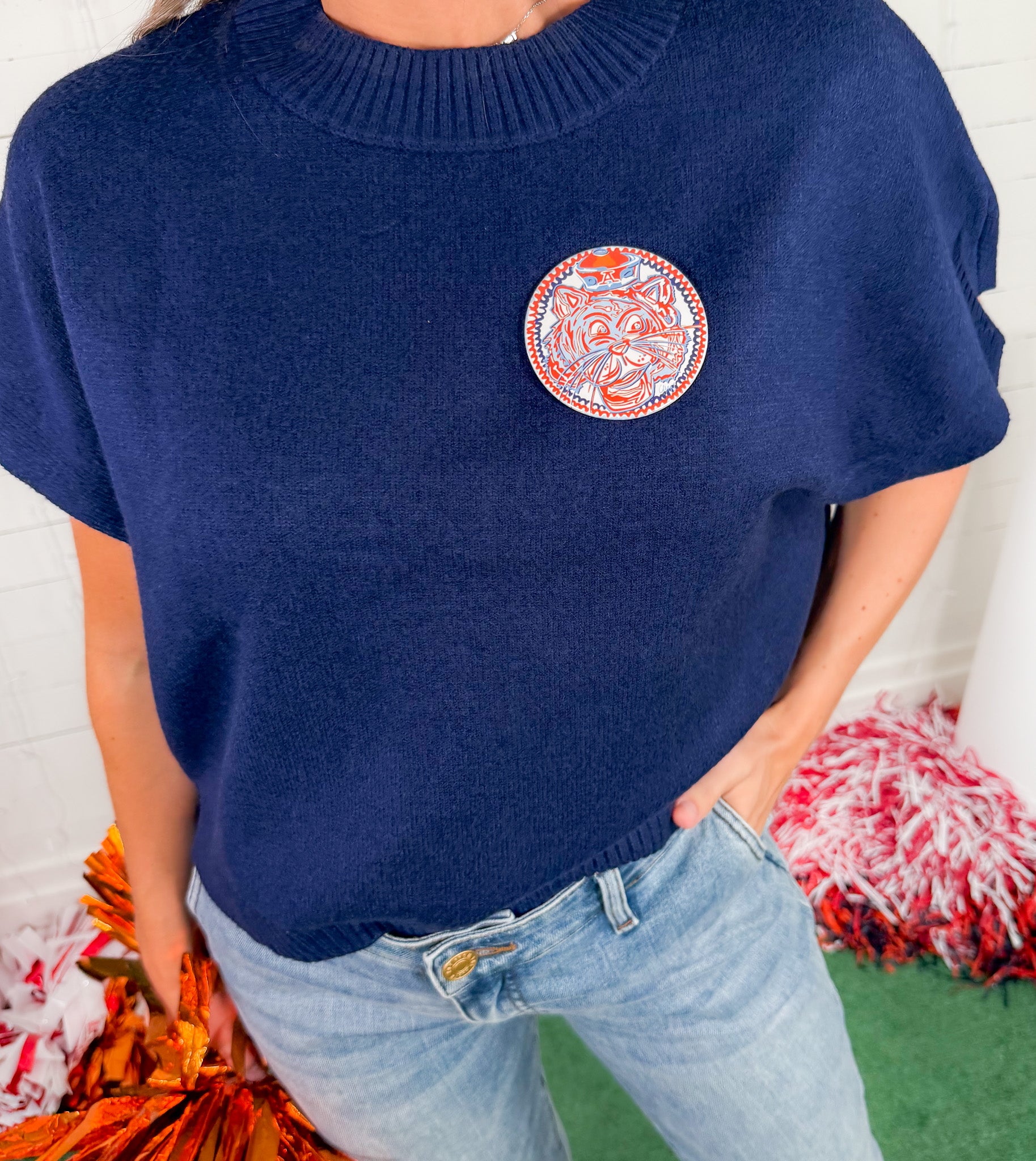 Person wearing a navy sweater with a circular orange and white design, standing against a white brick wall.