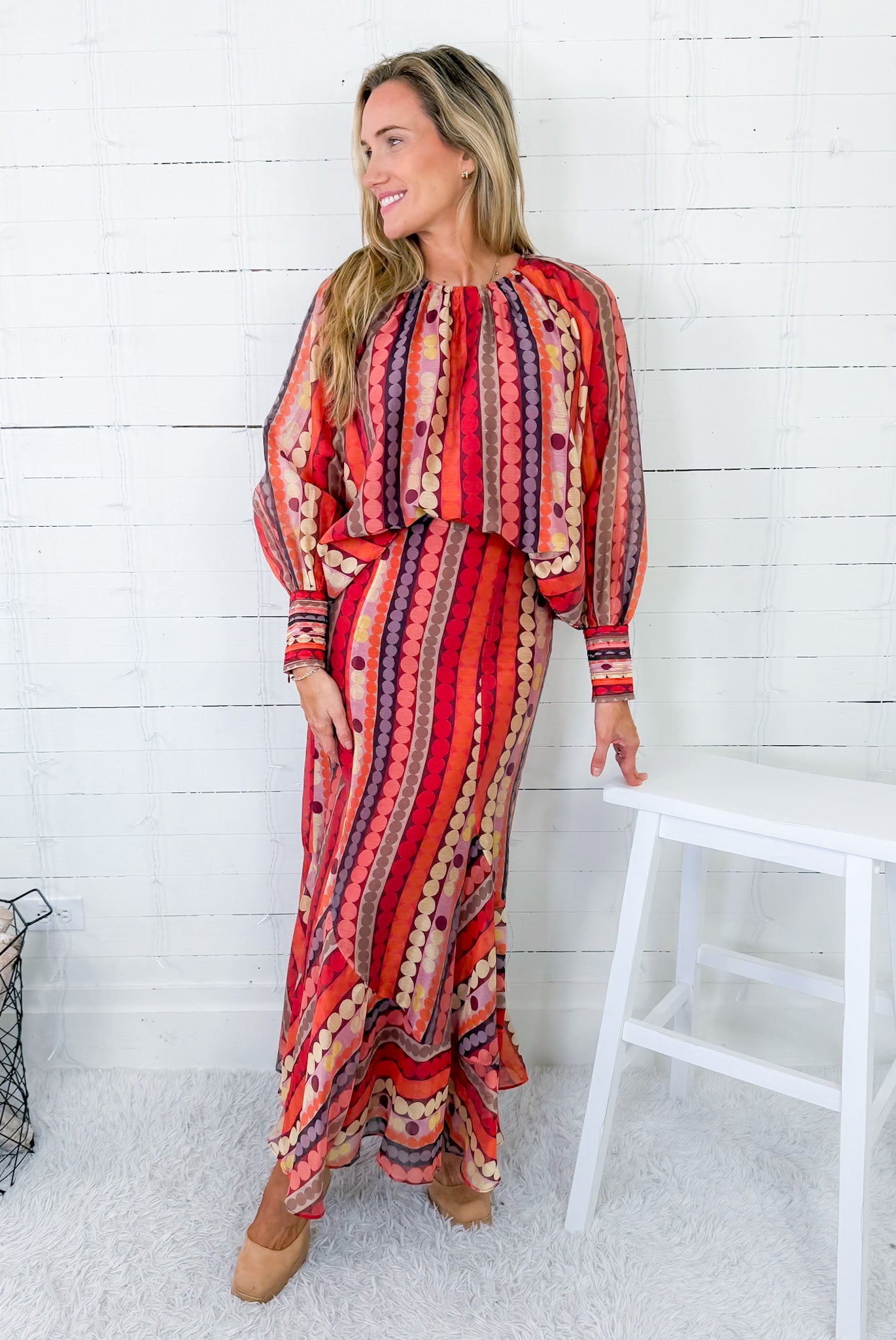 Woman wearing a red and orange patterned dress standing against a white wooden panel wall.