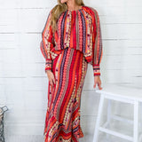 Woman wearing a red and orange patterned dress standing against a white wooden panel wall.