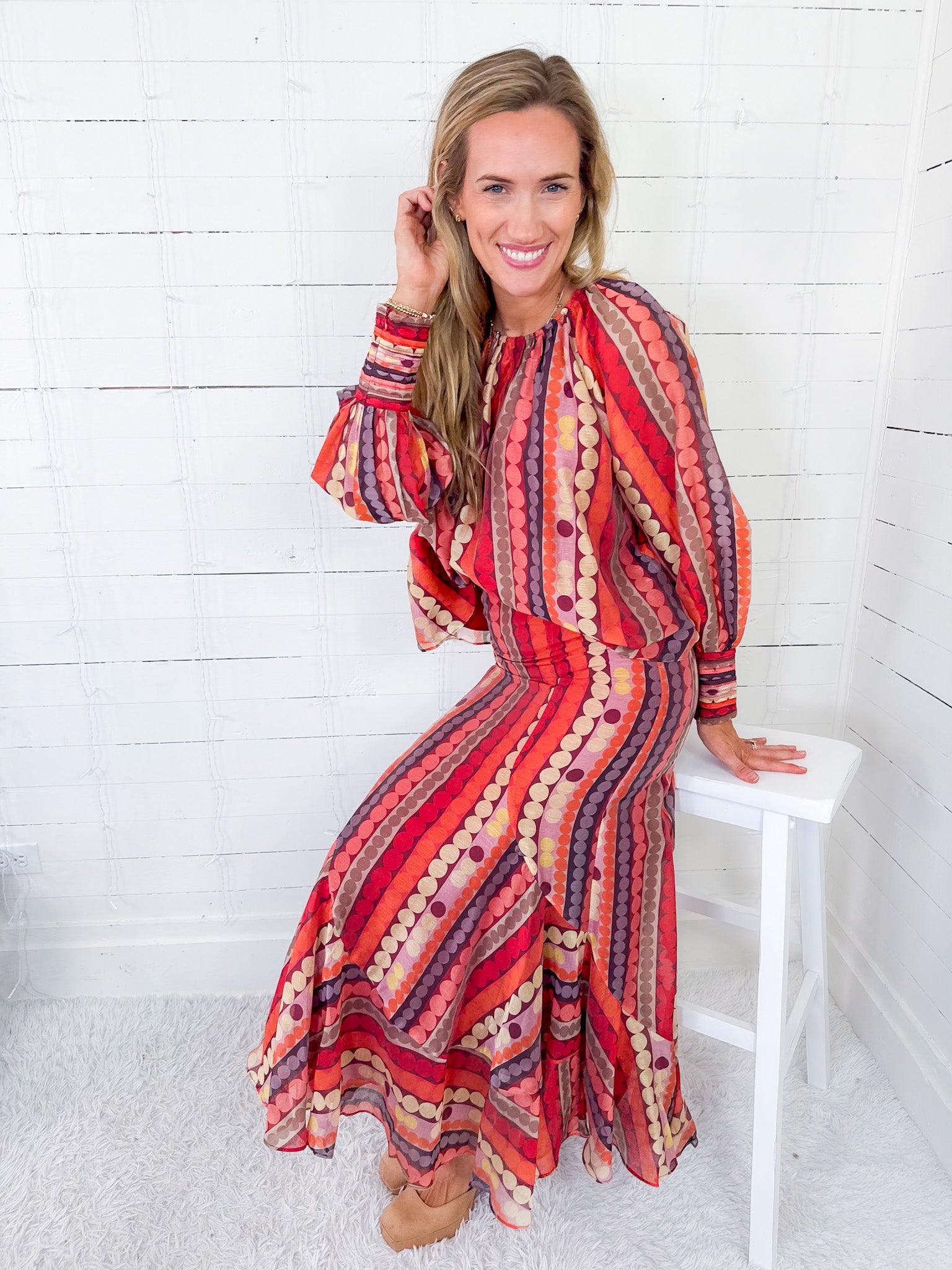 Woman wearing a colorful patterned dress sitting on a white stool against a white wooden panel background.