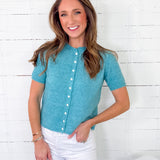 Woman wearing a teal button-up shirt and white pants against a white wooden wall.