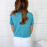 Person wearing a teal top and white pants against a white wooden panel background