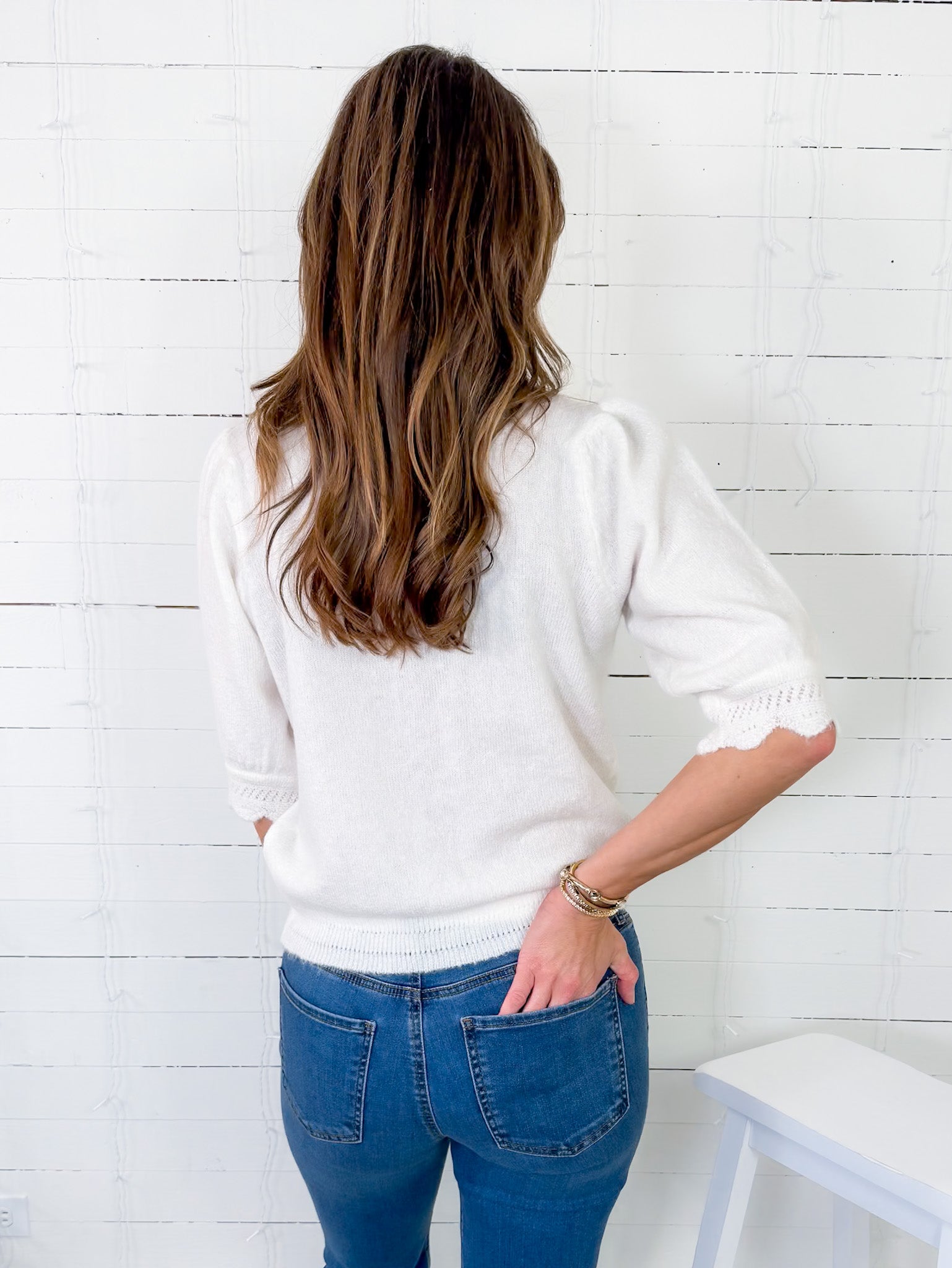 Person wearing a white sweater and blue jeans against a white wooden panel background