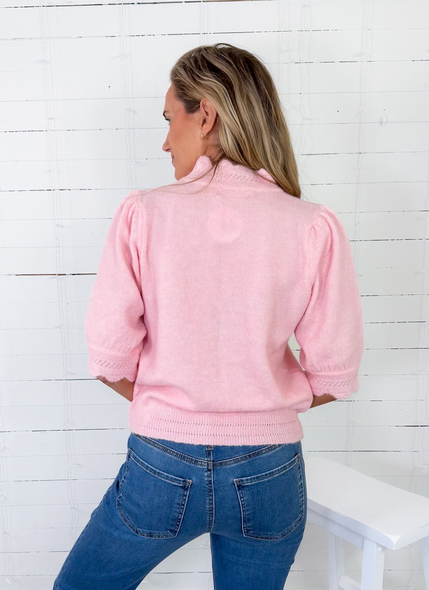 Woman wearing a pink jacket and blue jeans against a white wooden panel background