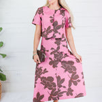 A woman modeling a pink short-sleeve midi dress with a large dark brown floral print, standing against a white wood-paneled wall.