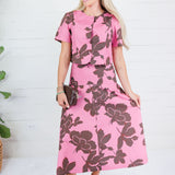 A woman modeling a pink short-sleeve midi dress with a large dark brown floral print, standing against a white wood-paneled wall.