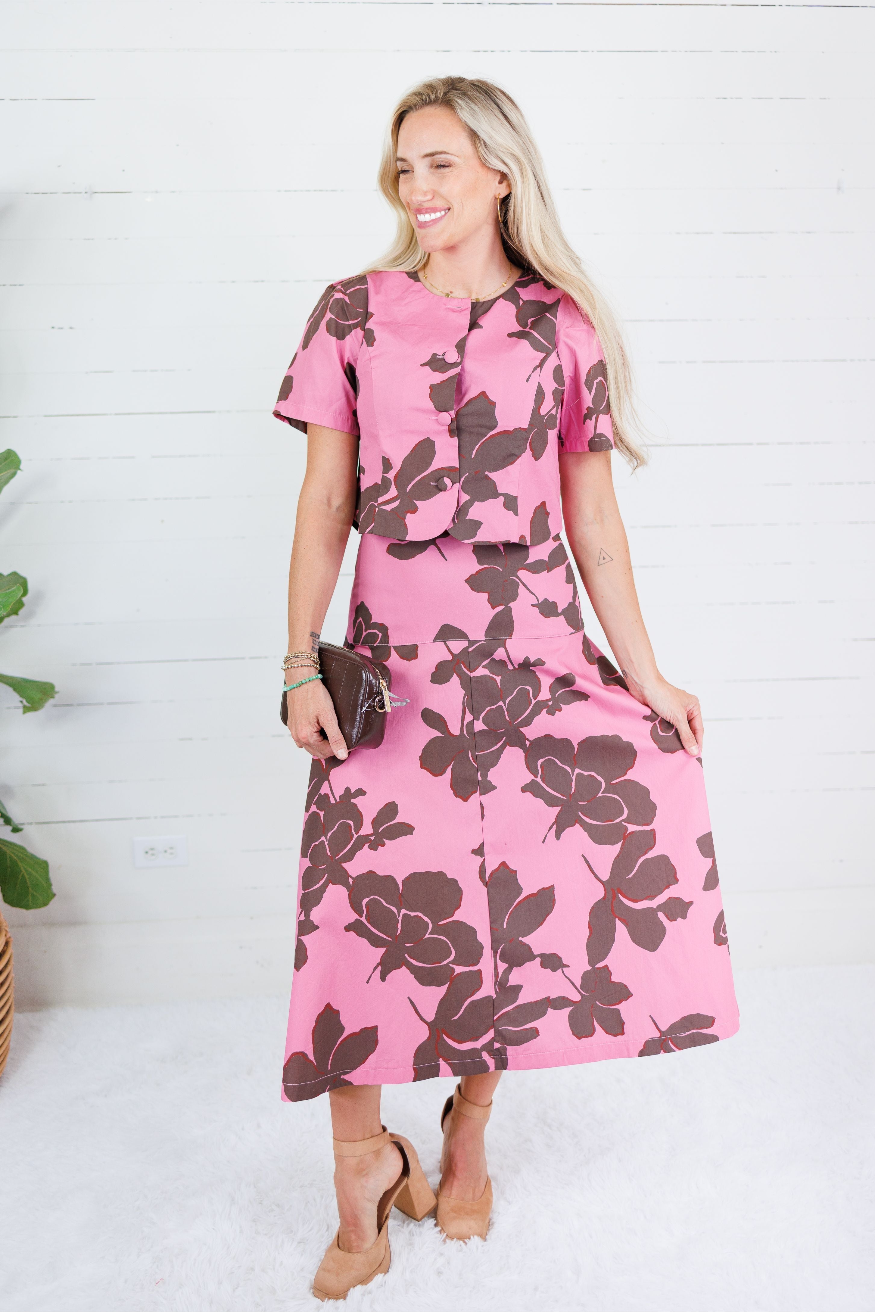 A woman modeling a pink short-sleeve midi dress with a large dark brown floral print, standing against a white wood-paneled wall.