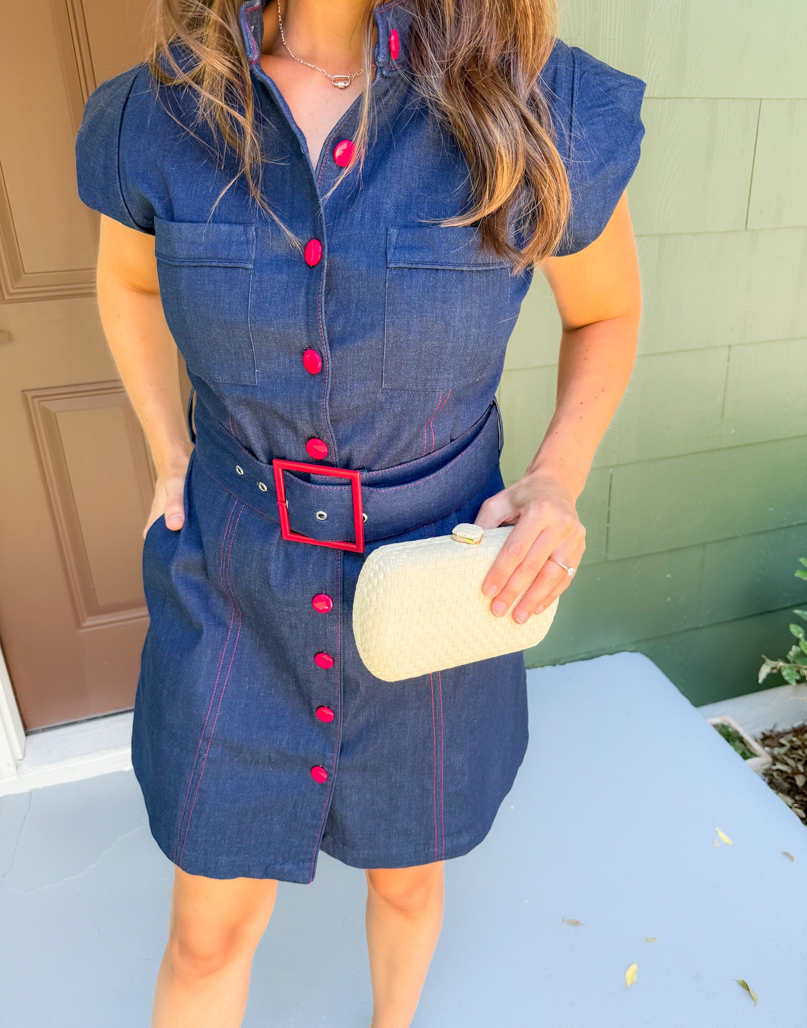 Woman wearing a denim dress with red buttons and a belt, holding a beige clutch.