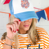 Person wearing a blue and white cap with a logo, holding it up to their face, against a colorful flag background.