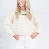 A smiling blonde woman wearing a cream and yellow striped long-sleeve knit top with white pants, standing against a white wood plank wall.