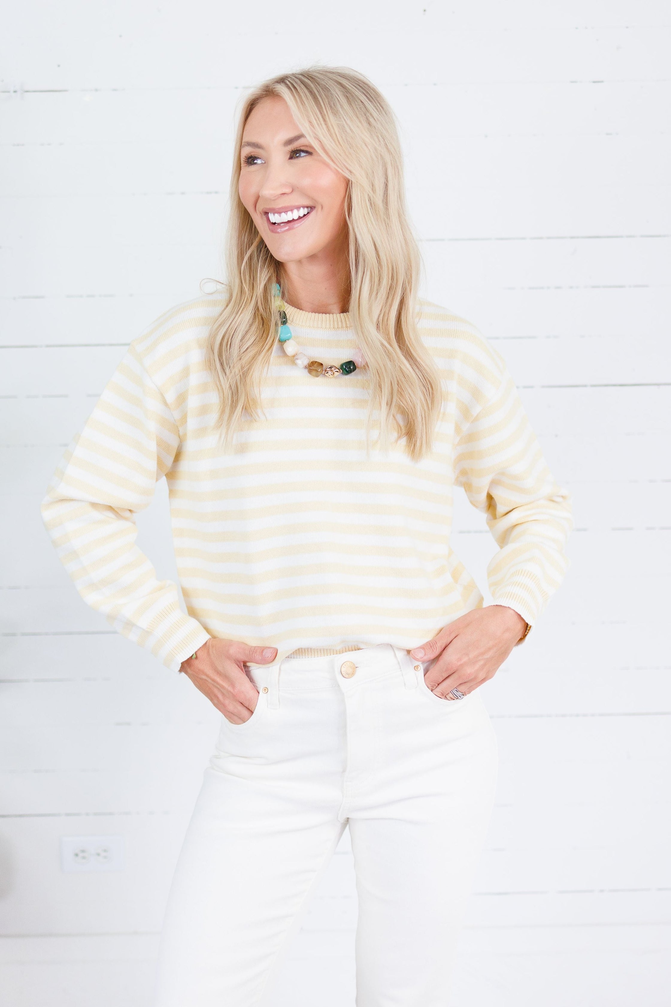 A smiling blonde woman wearing a cream and yellow striped long-sleeve knit top with white pants, standing against a white wood plank wall.