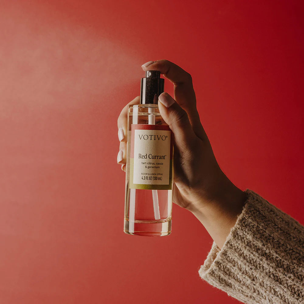 Hand holding a bottle of Votivo Red Currant body spray against a red background