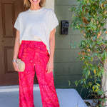 Woman wearing a white top and bright pink patterned pants standing outside a house.