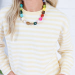 A woman wearing a cream and yellow striped knit sweater and a chunky multi-colored beaded necklace, shown from the chest up against a bright white background.