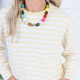 A woman wearing a cream and yellow striped knit sweater and a chunky multi-colored beaded necklace, shown from the chest up against a bright white background.
