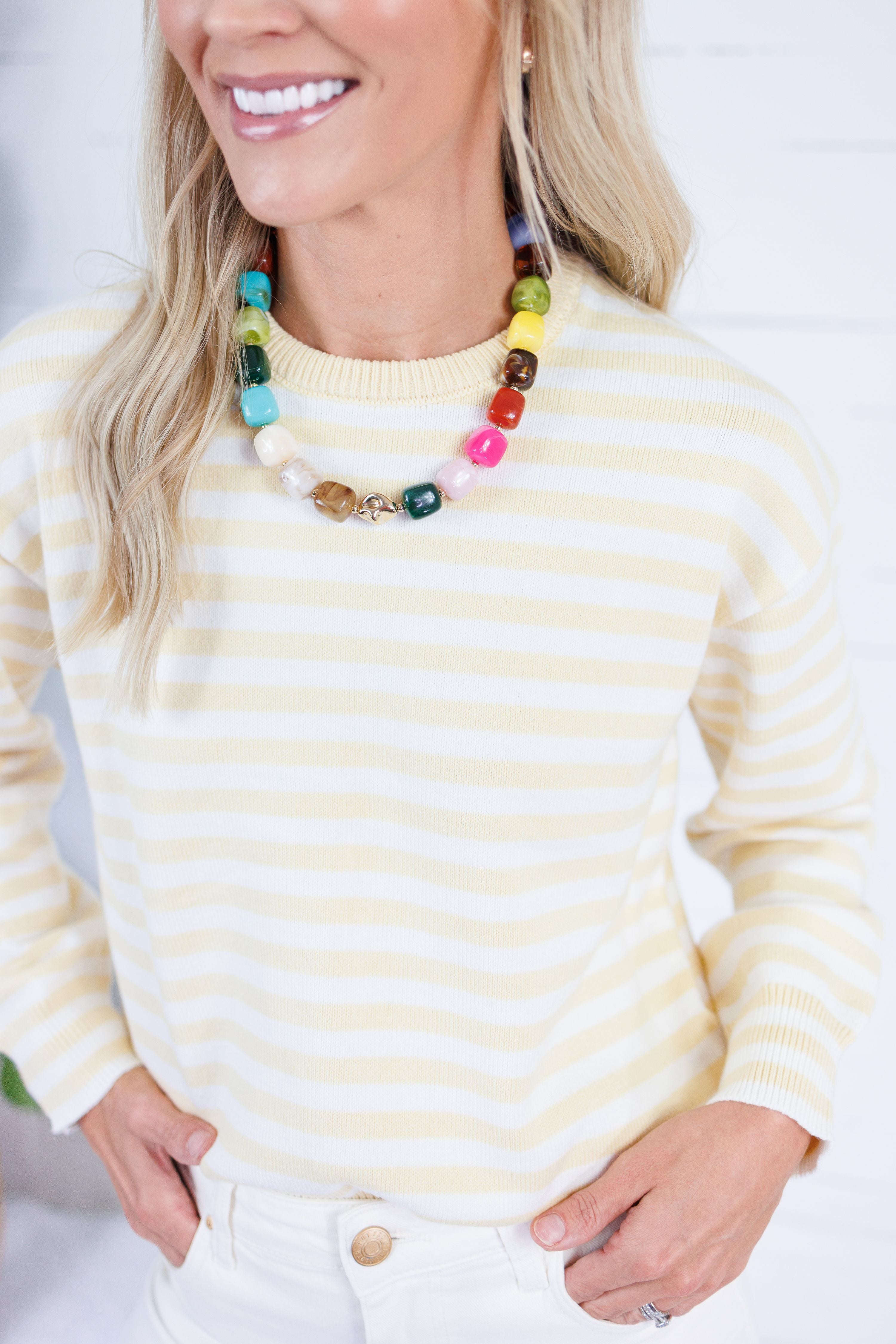 A woman wearing a cream and yellow striped knit sweater and a chunky multi-colored beaded necklace, shown from the chest up against a bright white background.
