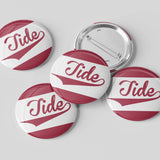 Four round pin-back buttons with a white and dark red design featuring the word 'Tide' in cursive, with one button showing the metal pin-back backing, on a light gray background.