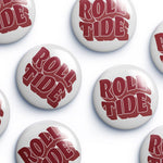 Several round white button badges with the words "ROLL TIDE" in large red stylized lettering, arranged on a light gray background.