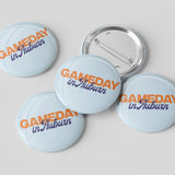 Five round pin-back buttons with a light blue background and the text 'GAMEDAY in Auburn' in orange and navy blue, with one button showing the silver metal pin-back mechanism, on a light gray background.