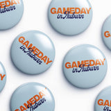 A repeating pattern of round light blue buttons with the text 'GAMEDAY in Auburn' printed in orange and dark blue, arranged on a white background.