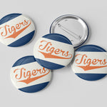 Five round pin-back buttons with a vintage sports design featuring the word 'Tigers' in orange script over diagonal navy and white stripes, shown on a light gray background with one button facing up and four facing down to show the metal pin-back mechanism.
