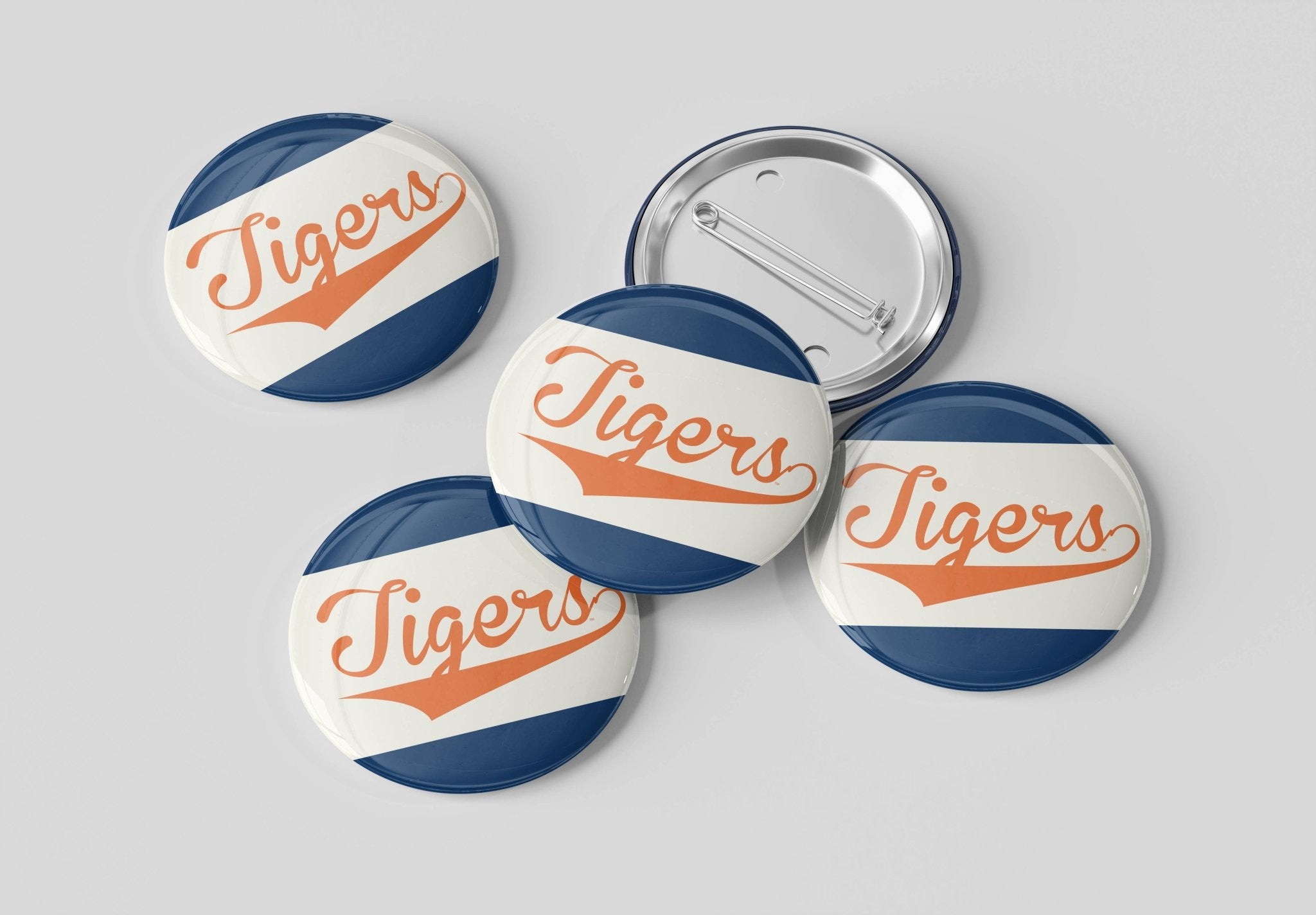 Five round pin-back buttons with a vintage sports design featuring the word 'Tigers' in orange script over diagonal navy and white stripes, shown on a light gray background with one button facing up and four facing down to show the metal pin-back mechanism.