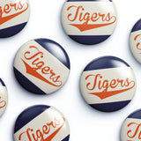 Several round glossy buttons with the word "Tigers" in orange script over diagonal navy and off-white stripes, arranged on a white background.