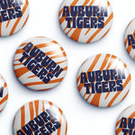 Several round button badges with the Auburn Tigers logo, featuring orange and white tiger stripes and navy blue lettering, arranged on a white background.