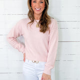 Woman wearing a light pink sweater and white pants against a white wooden panel background