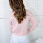 Person wearing a light pink sweater and white pants against a white wooden panel background