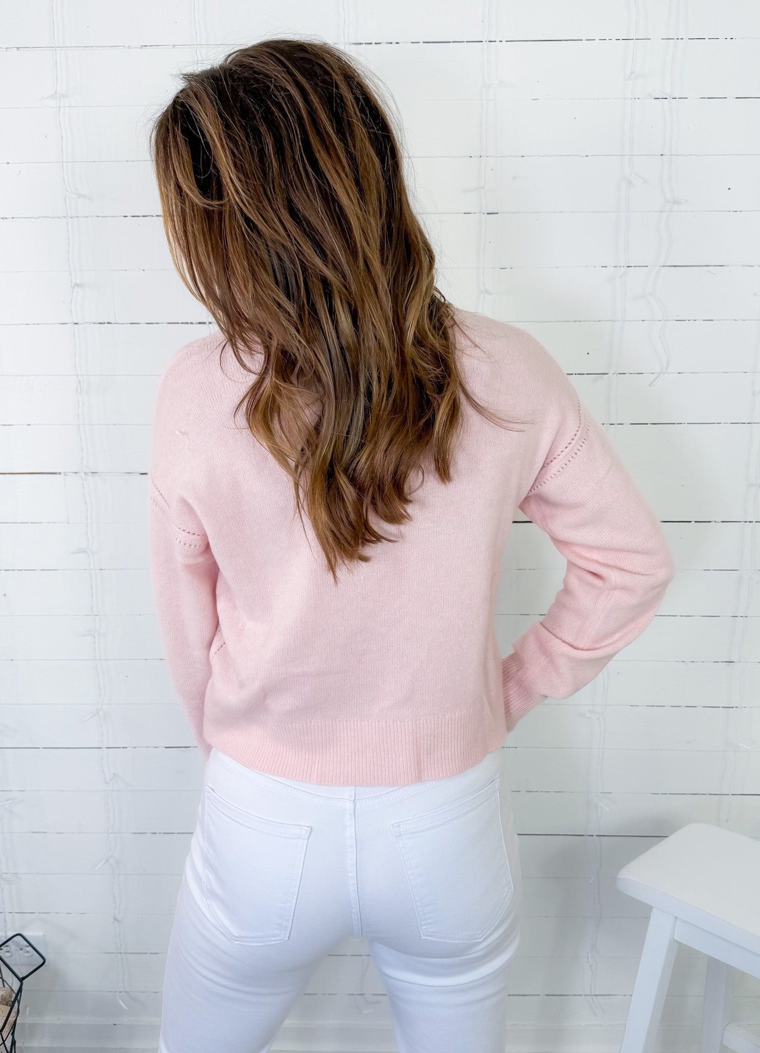Person wearing a light pink sweater and white pants against a white wooden panel background