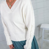 Person wearing a white sweater with long sleeves and a V-neckline, standing against a light-colored wall.
