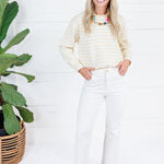 A woman stands in a bright white room wearing a cream and yellow striped long-sleeve knit top, white pants, and tan flats. A large green potted plant is on the left side of the frame.