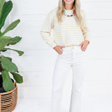 A woman stands in a bright white room wearing a cream and yellow striped long-sleeve knit top, white pants, and tan flats. A large green potted plant is on the left side of the frame.