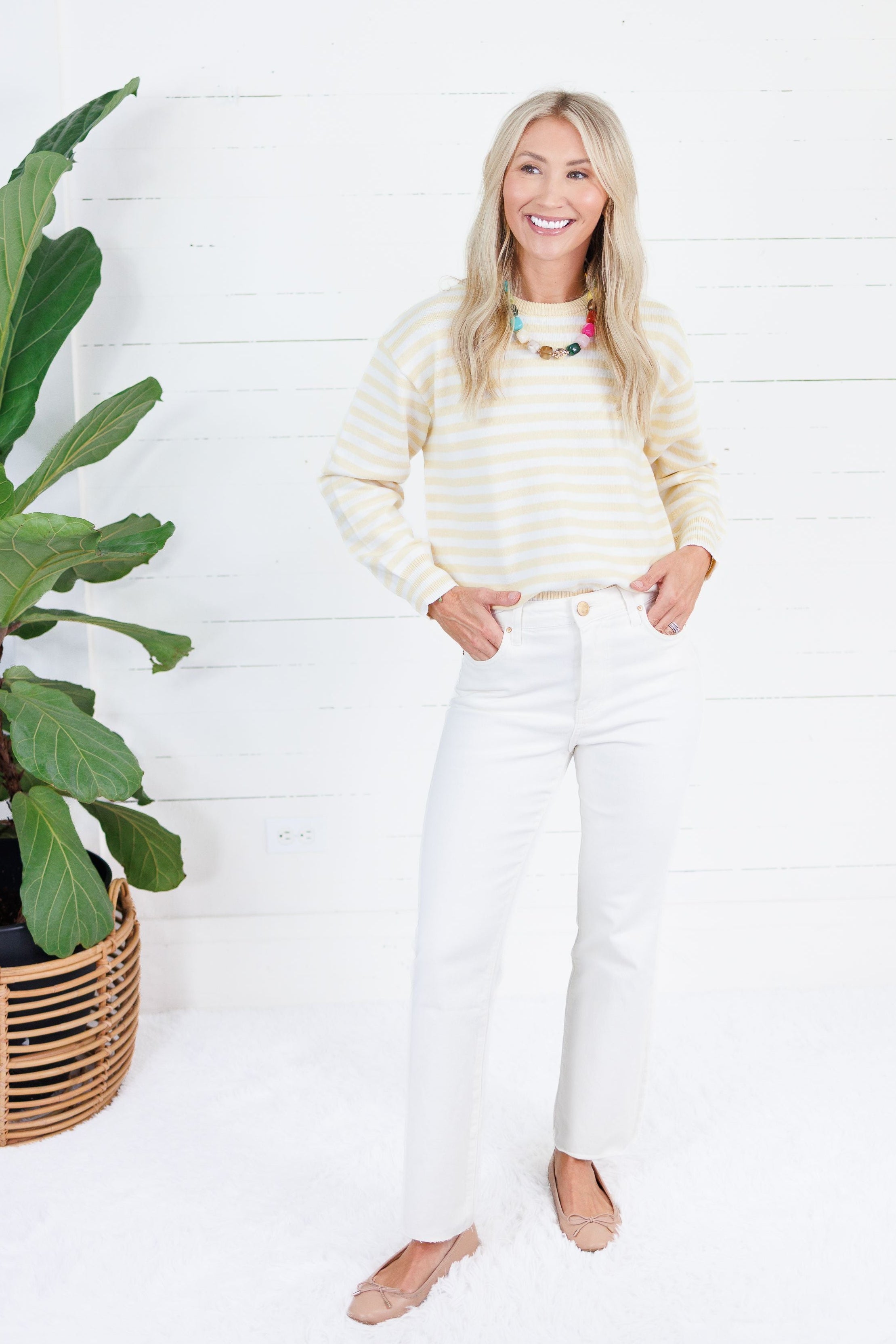 A woman stands in a bright white room wearing a cream and yellow striped long-sleeve knit top, white pants, and tan flats. A large green potted plant is on the left side of the frame.