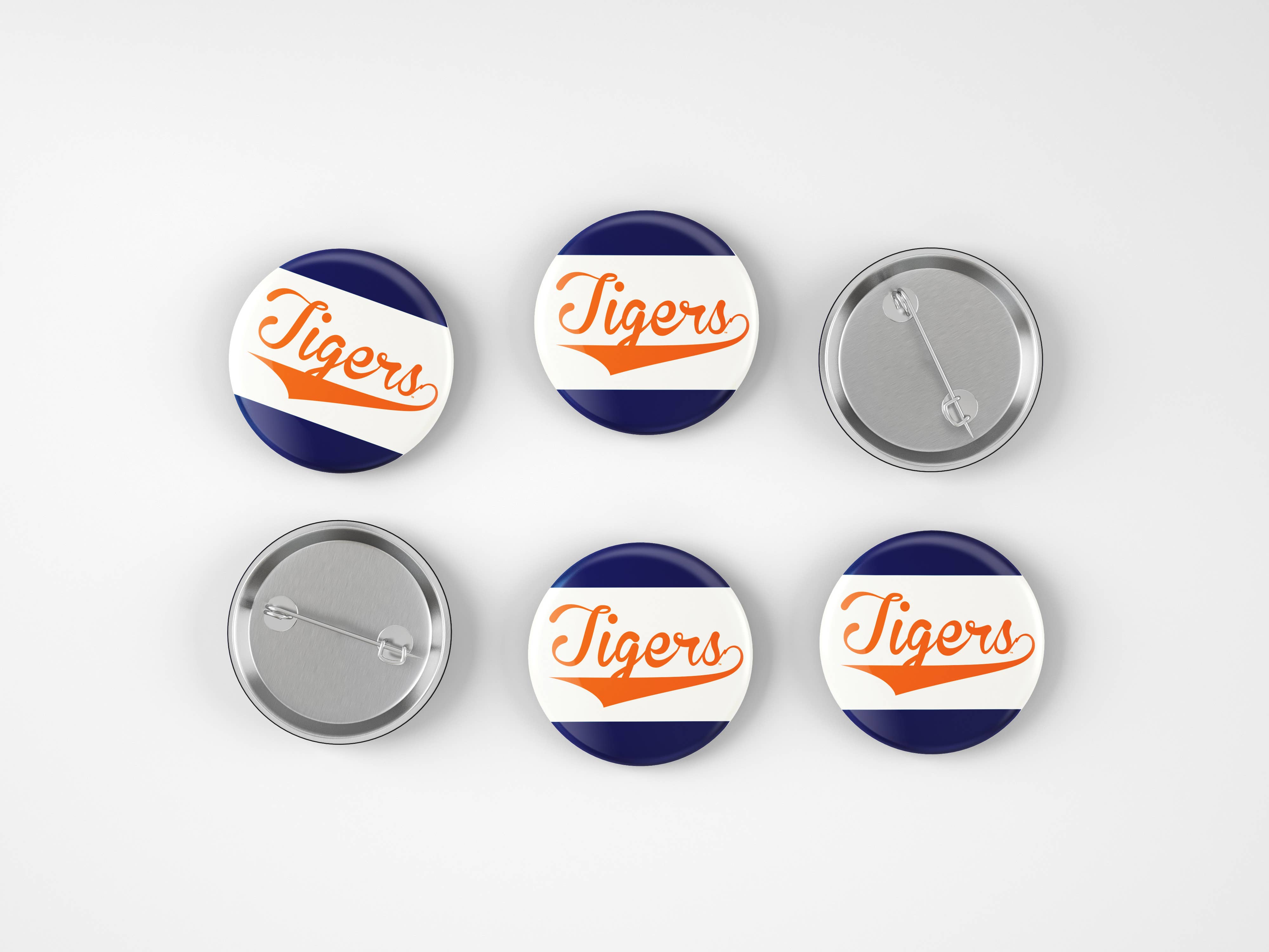 Six round pin-back buttons on a light gray background, showing both the front and back views. The front faces feature a navy blue and white diagonal striped design with the word 'Tigers' in orange script.