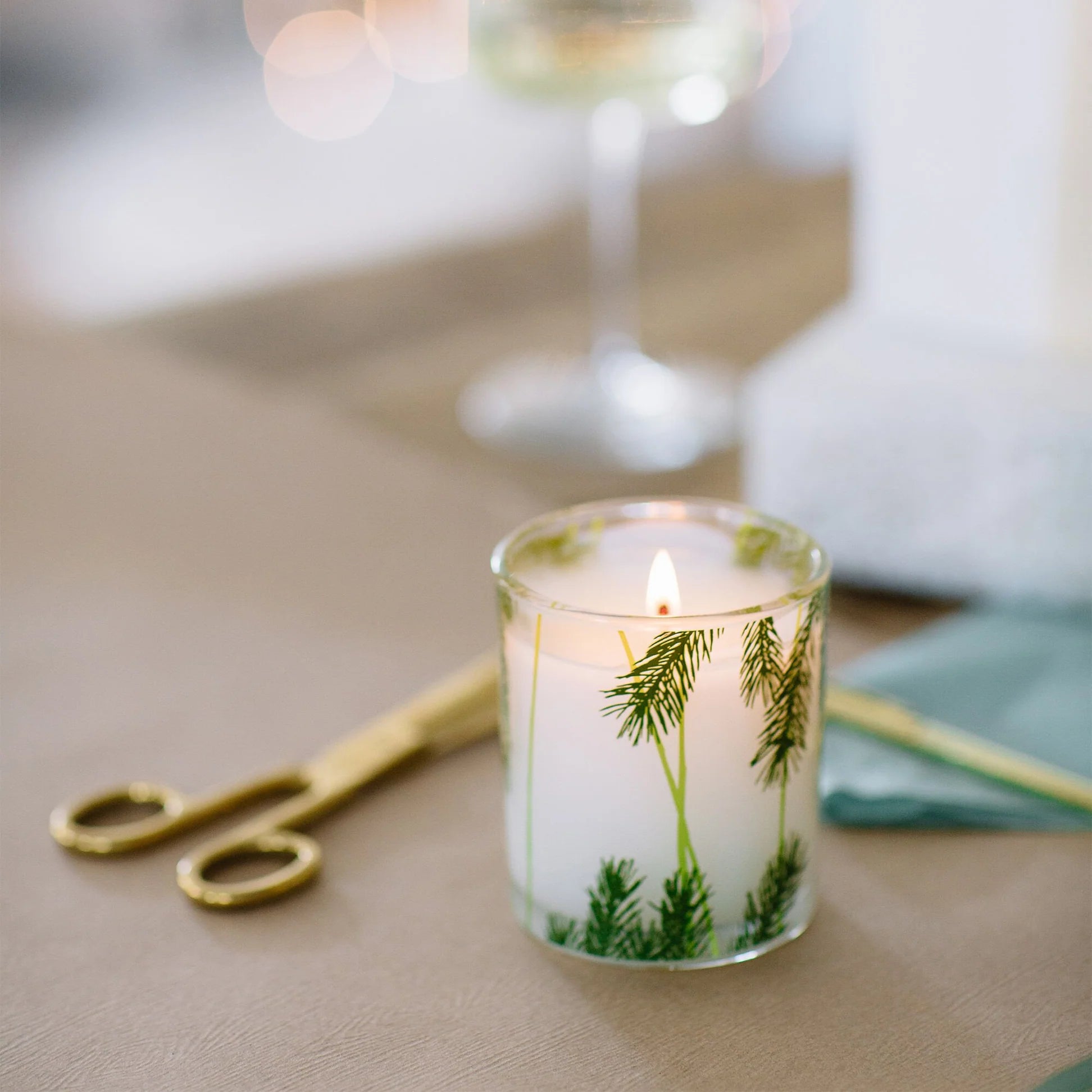 Candle with greenery design on a table with scissors and a glass in the background