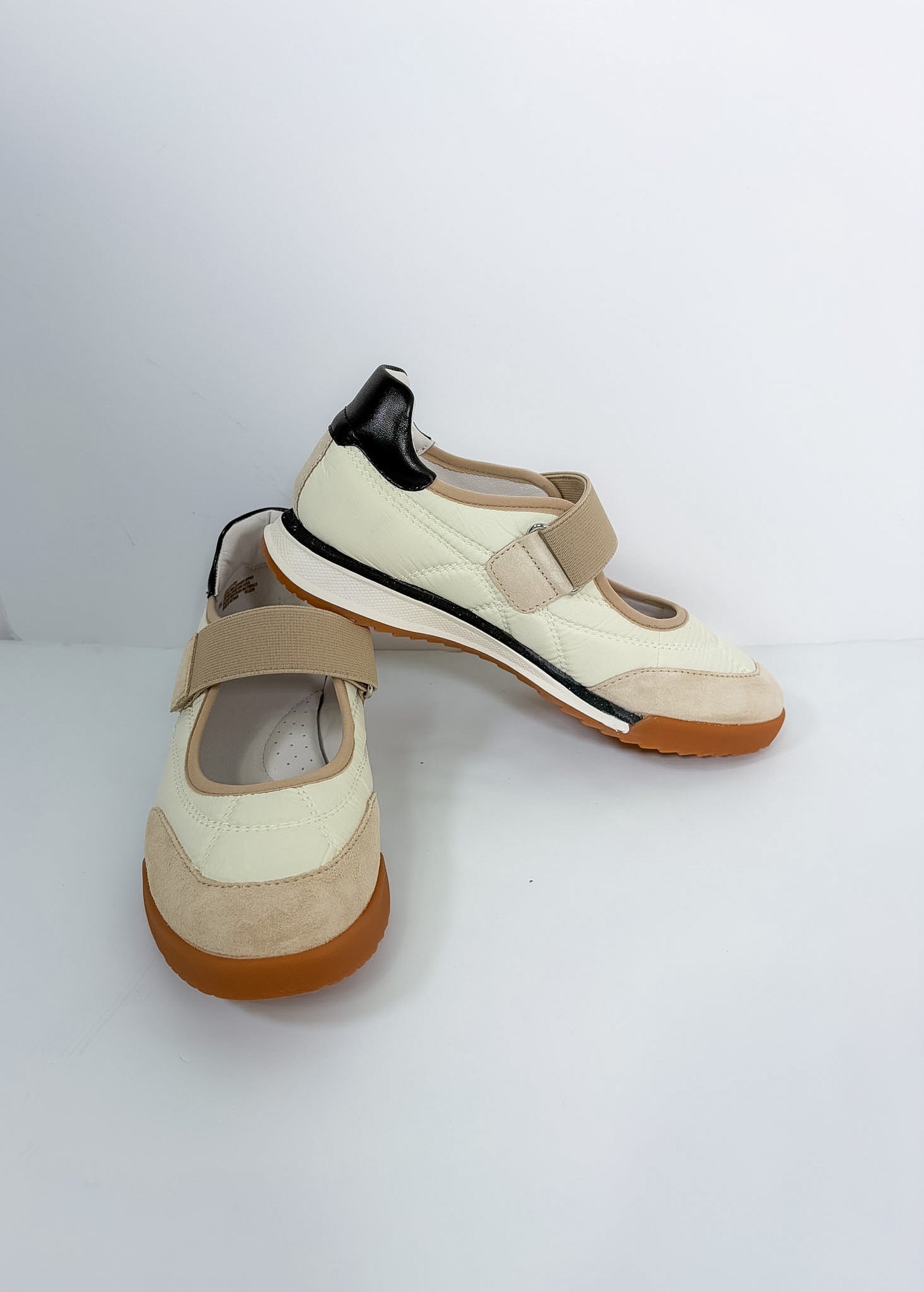 A pair of women's Mary Jane style sneakers in cream and beige suede, featuring a beige hook-and-loop strap, a black heel accent, and a brown sole.