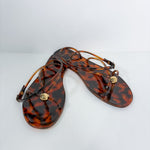 A pair of women's thong sandals in a glossy amber-brown tortoiseshell pattern, featuring thin wrap-around straps and a small gold-tone circular medallion on the toe post, displayed on a clean white background.