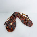 A pair of women's thong sandals in a glossy amber-brown tortoiseshell pattern, featuring thin wrap-around straps and a small gold-tone circular medallion on the toe post, displayed on a clean white background.