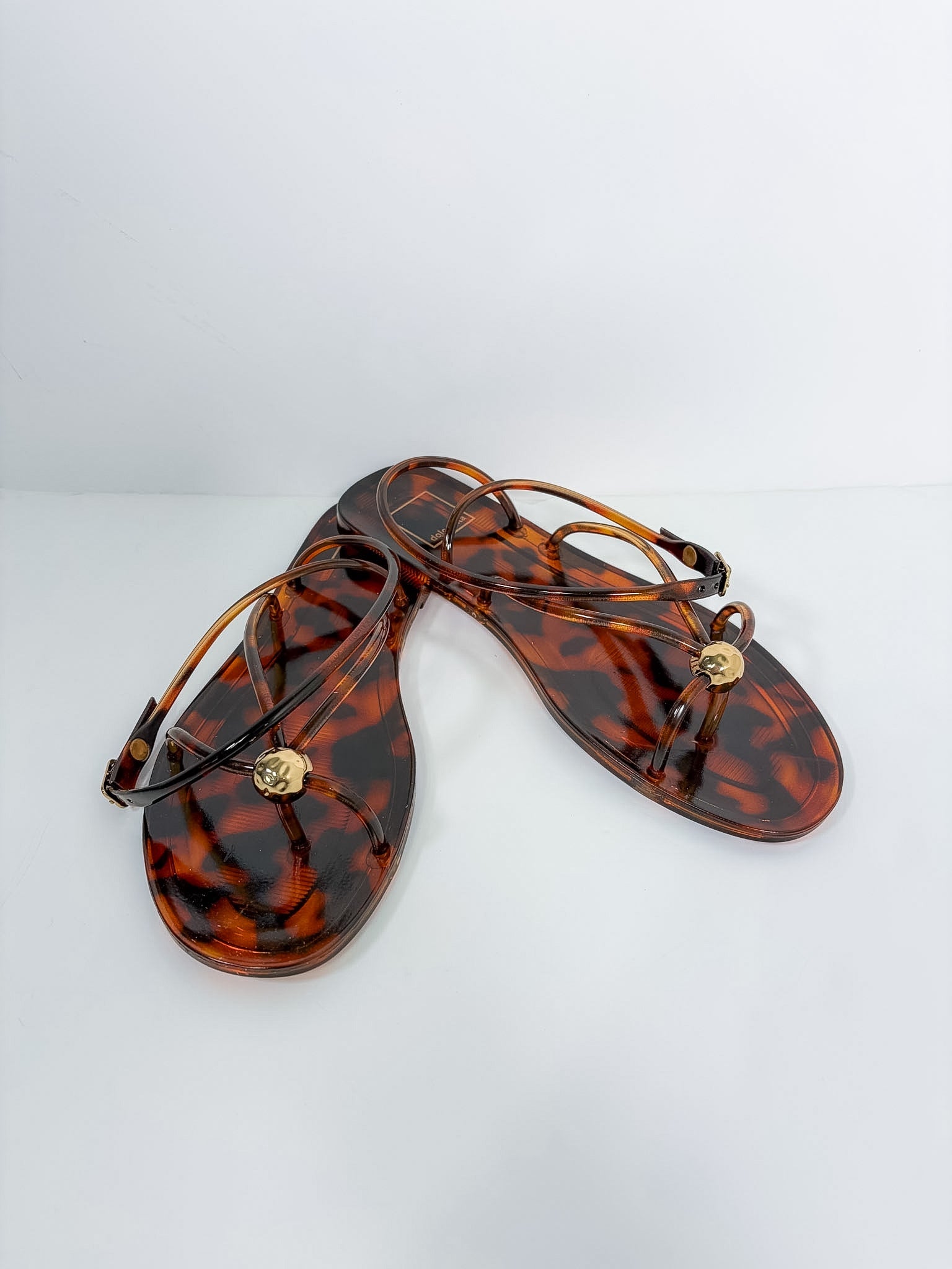 A pair of women's thong sandals in a glossy amber-brown tortoiseshell pattern, featuring thin wrap-around straps and a small gold-tone circular medallion on the toe post, displayed on a clean white background.