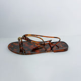 A single flat thong sandal with a glossy tortoiseshell pattern in brown and amber, featuring thin wrap-around straps and a small gold-tone metal ring detail at the front, shown against a plain light gray background.