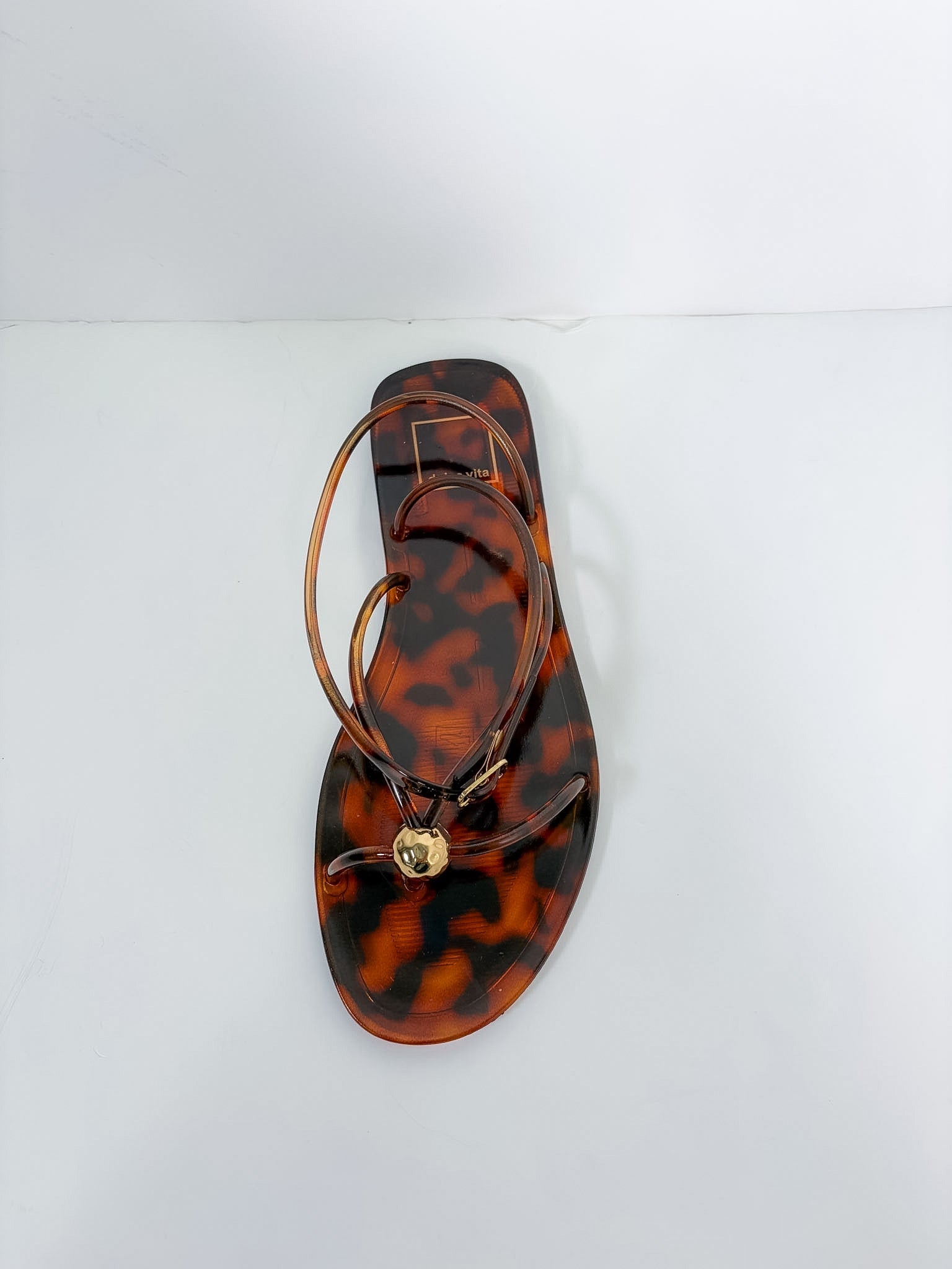 A single flat thong sandal with a glossy tortoiseshell pattern in brown and amber, featuring thin brown straps and a small gold-tone circular charm, photographed from a top-down angle on a white background.