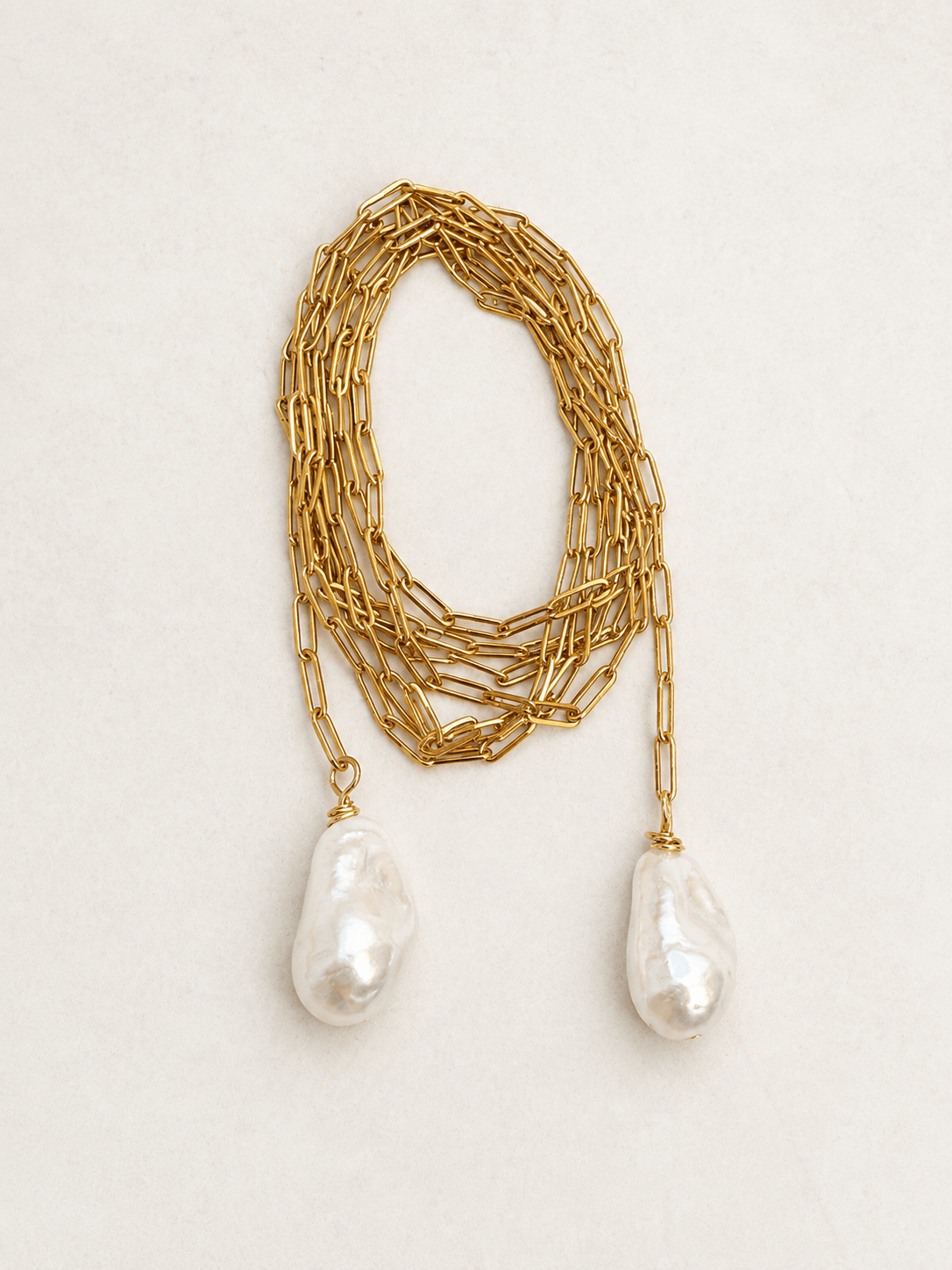 A gold chain necklace featuring two large, irregular white baroque pearls hanging from the ends, displayed on a light beige textured background.