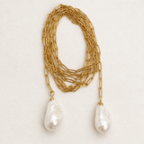 A gold chain necklace featuring two large, irregular white baroque pearls hanging from the ends, displayed on a light beige textured background.